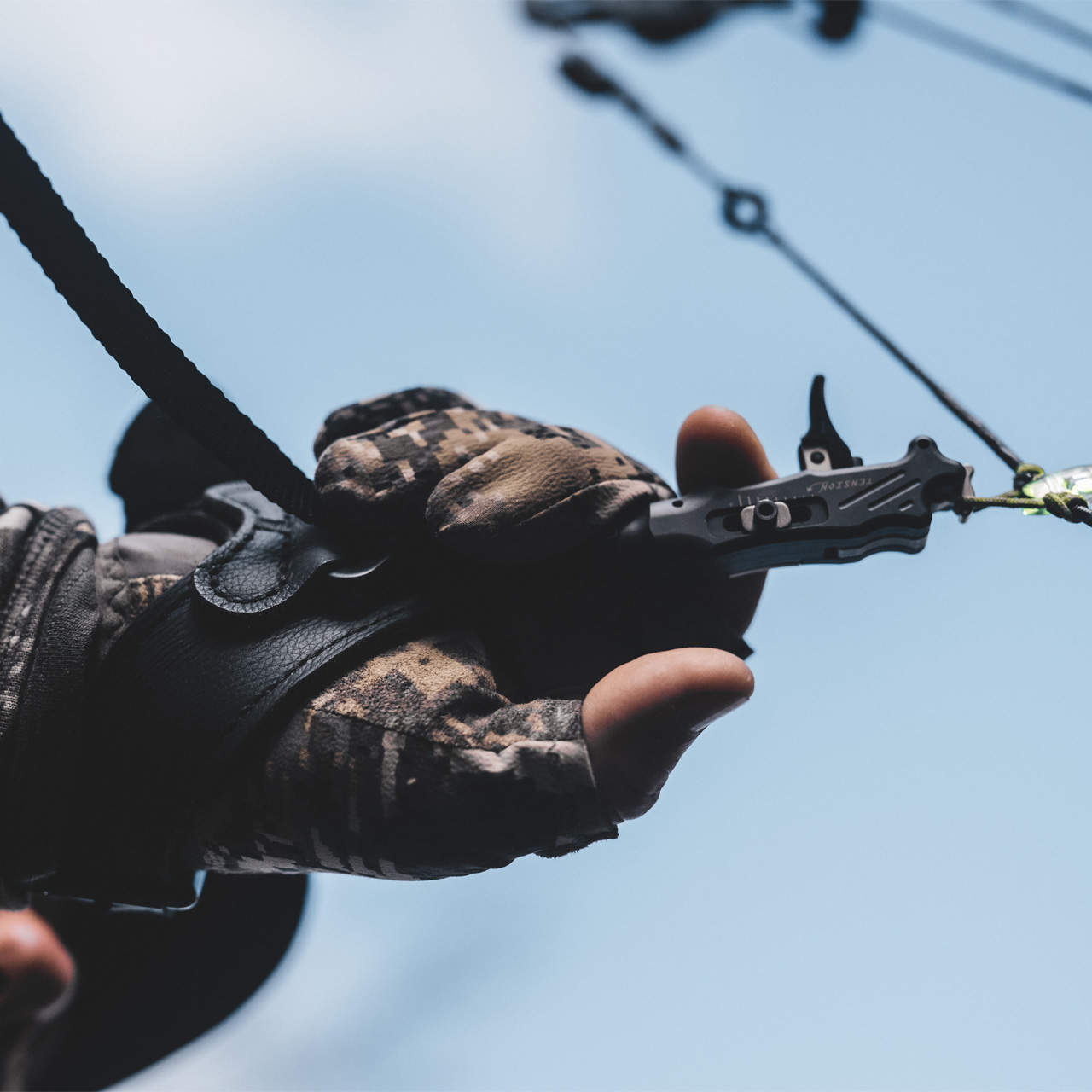 High-quality archery release held by a hunter wearing camouflage gloves against a blue sky.