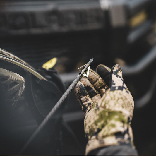 Camouflage glove holding arrow on bow within a hunting vehicle interior.