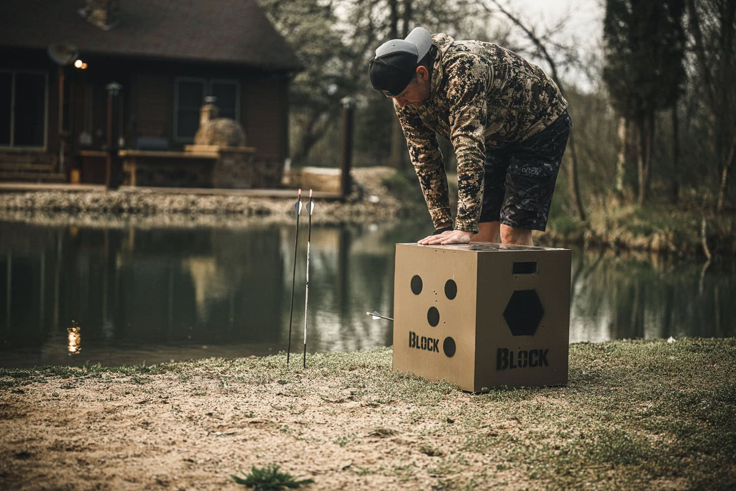 Archer practicing at water's edge with target block and arrows nearby in outdoor setting.