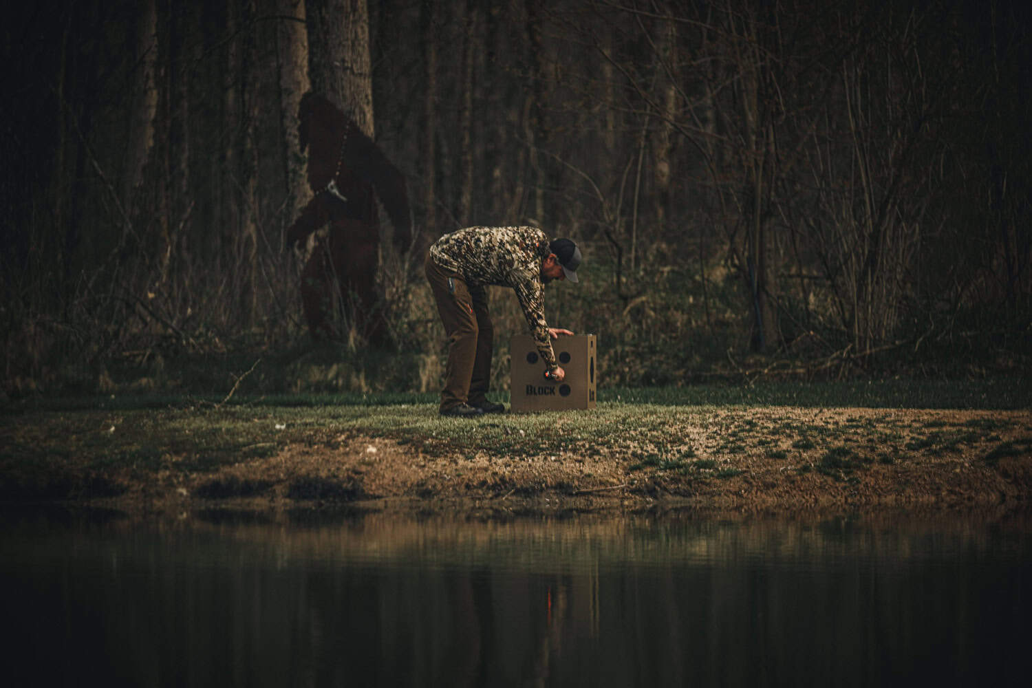 1. Man setting up a bowhunting target by a pond in the woods at dusk.