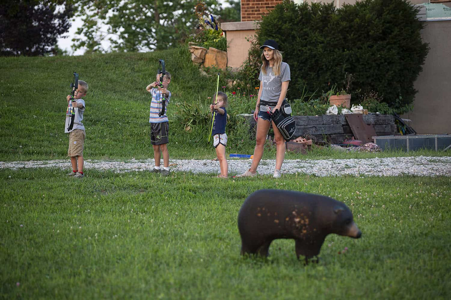 1. Children practicing archery outdoors with bows, supervised by a woman, in a grassy yard setting.