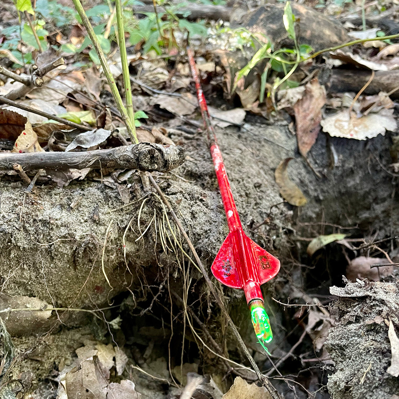 Bright red toy arrow with green flickering LED tip, resting on dirt and leaves in a nature setting.