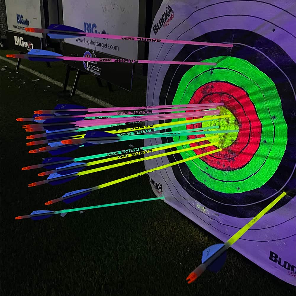 Brightly colored archery arrows shot at neon-lit target with concentric rings, illuminated at night.
