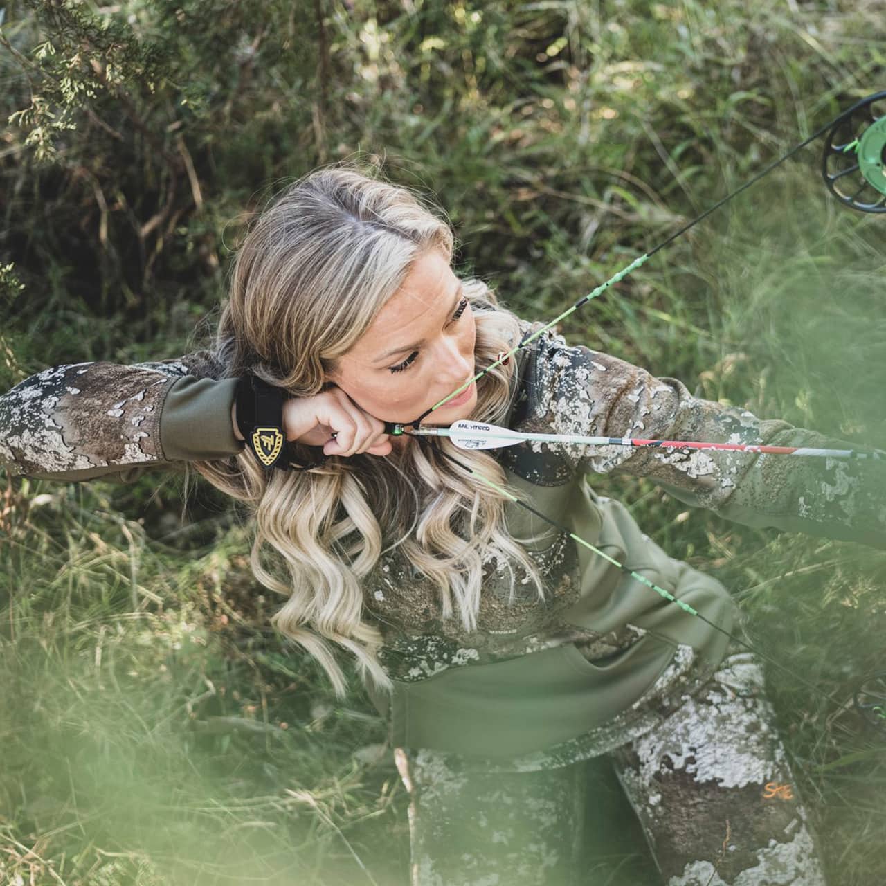 Female archer aiming her bow in a wooded outdoor setting.