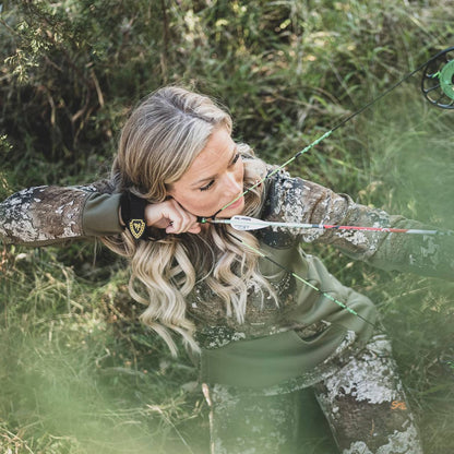 Female archer aiming her bow in a wooded outdoor setting.