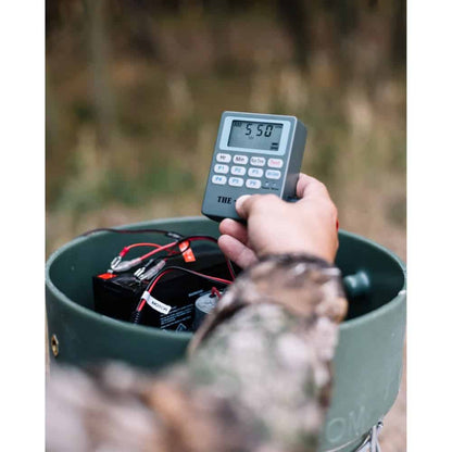 Hand holding a digital device measuring sound levels outdoors with a camo container.